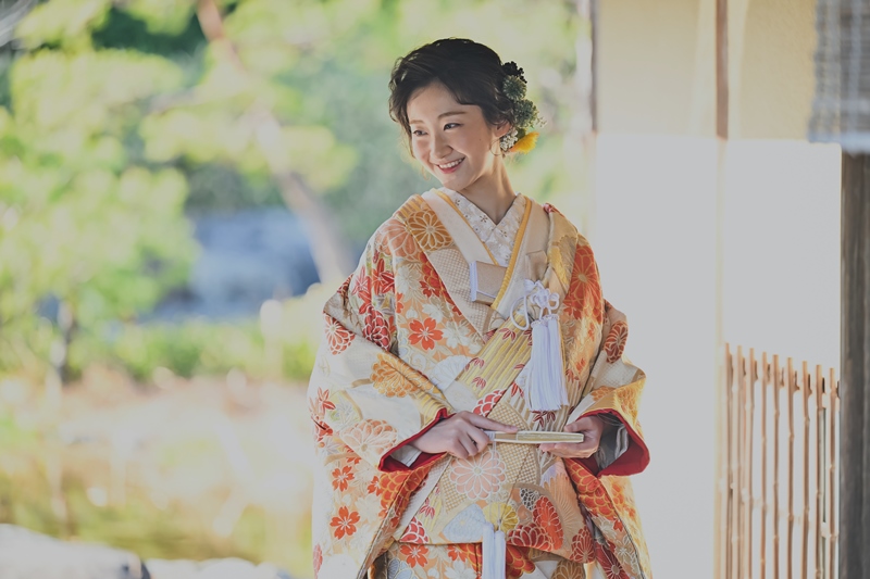 縁側で微笑む新婦、鮮やかな色打掛姿で扇を手に。