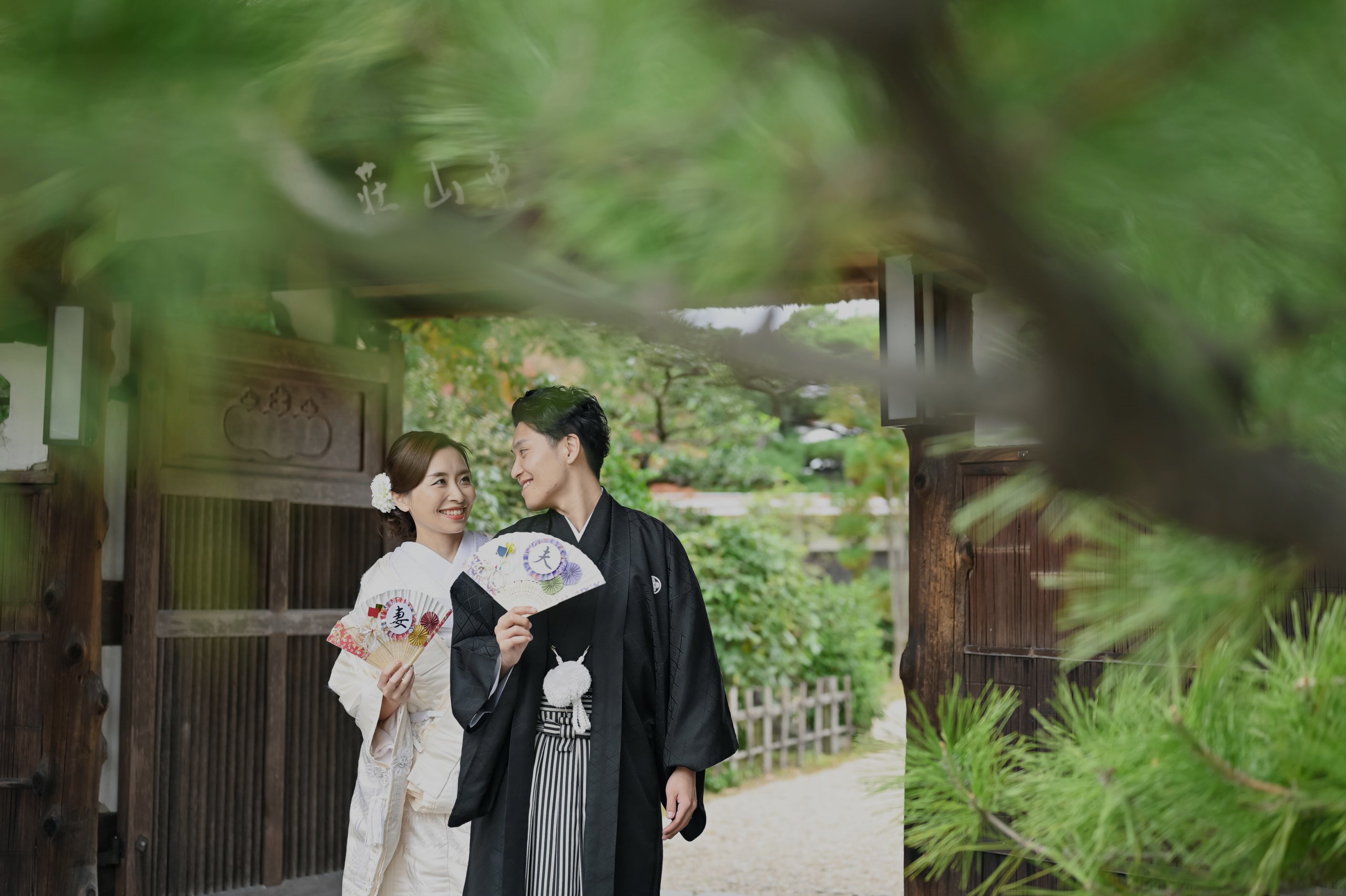 東山荘の門を背景に、末広扇を持ち見つめ合う新郎新婦、和装姿。