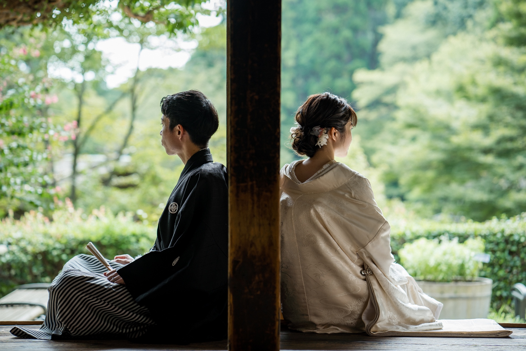 縁側で背中合わせに座り、庭を眺める新郎新婦、和装姿。