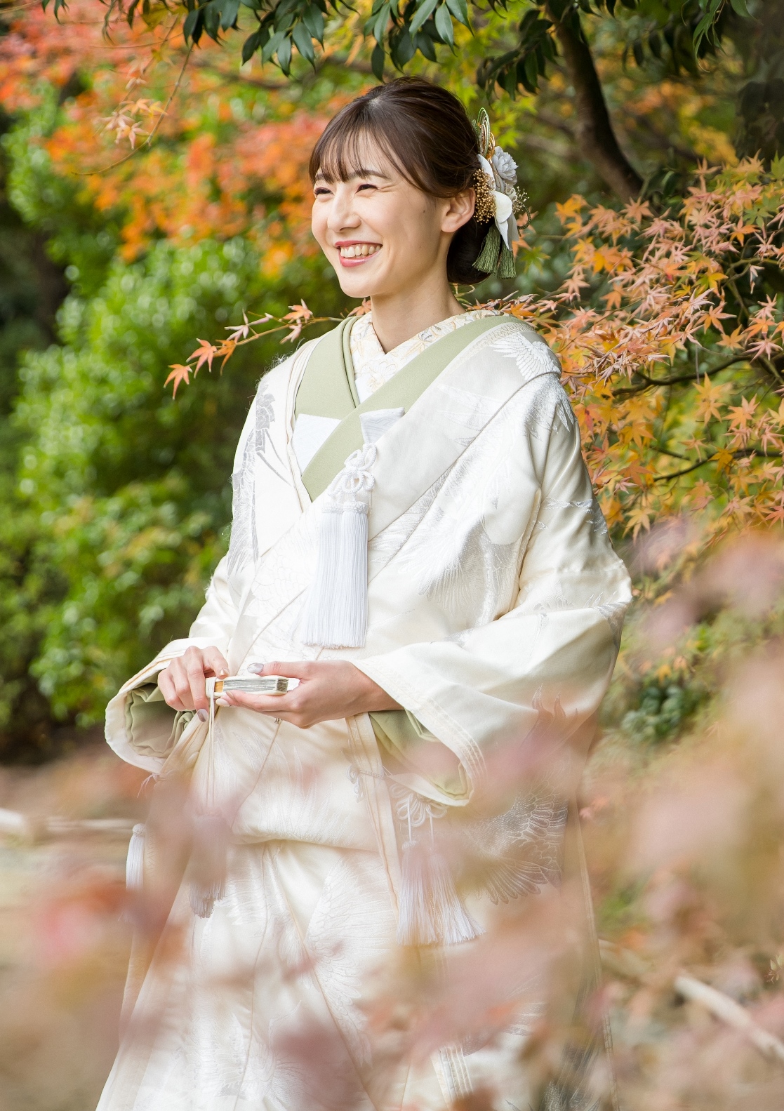 白無垢に緑の帯を合わせた新婦が、紅葉の中で明るく微笑む和装ポートレート。