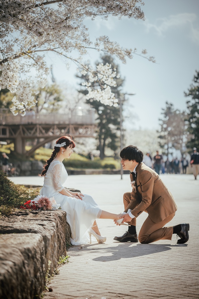 満開の桜の下、石垣に腰掛けた新婦の足元に跪いて靴を履かせる新郎の微笑ましいシーン。