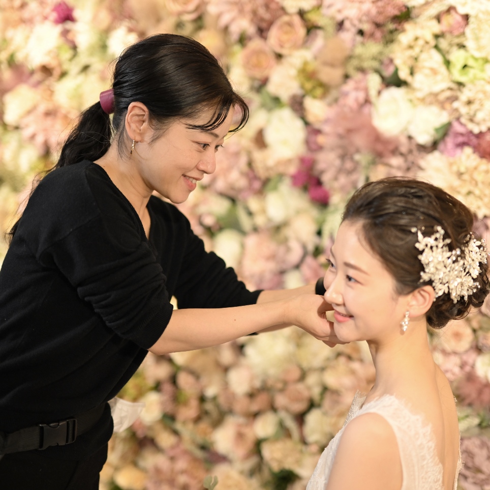 花の壁を背景に、新婦の顔にメイクを施すヘアメイクスタッフ多田。