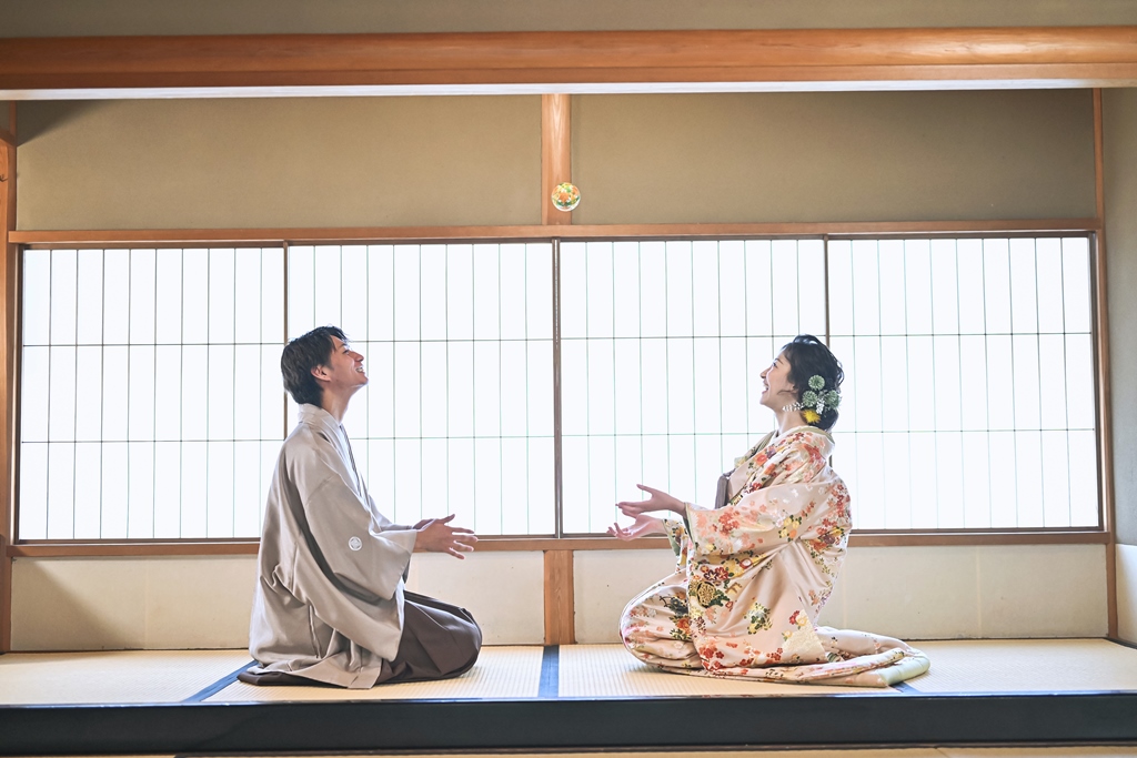 障子の間で紙風船を投げ合って笑う新郎新婦、色打掛と羽織袴姿。