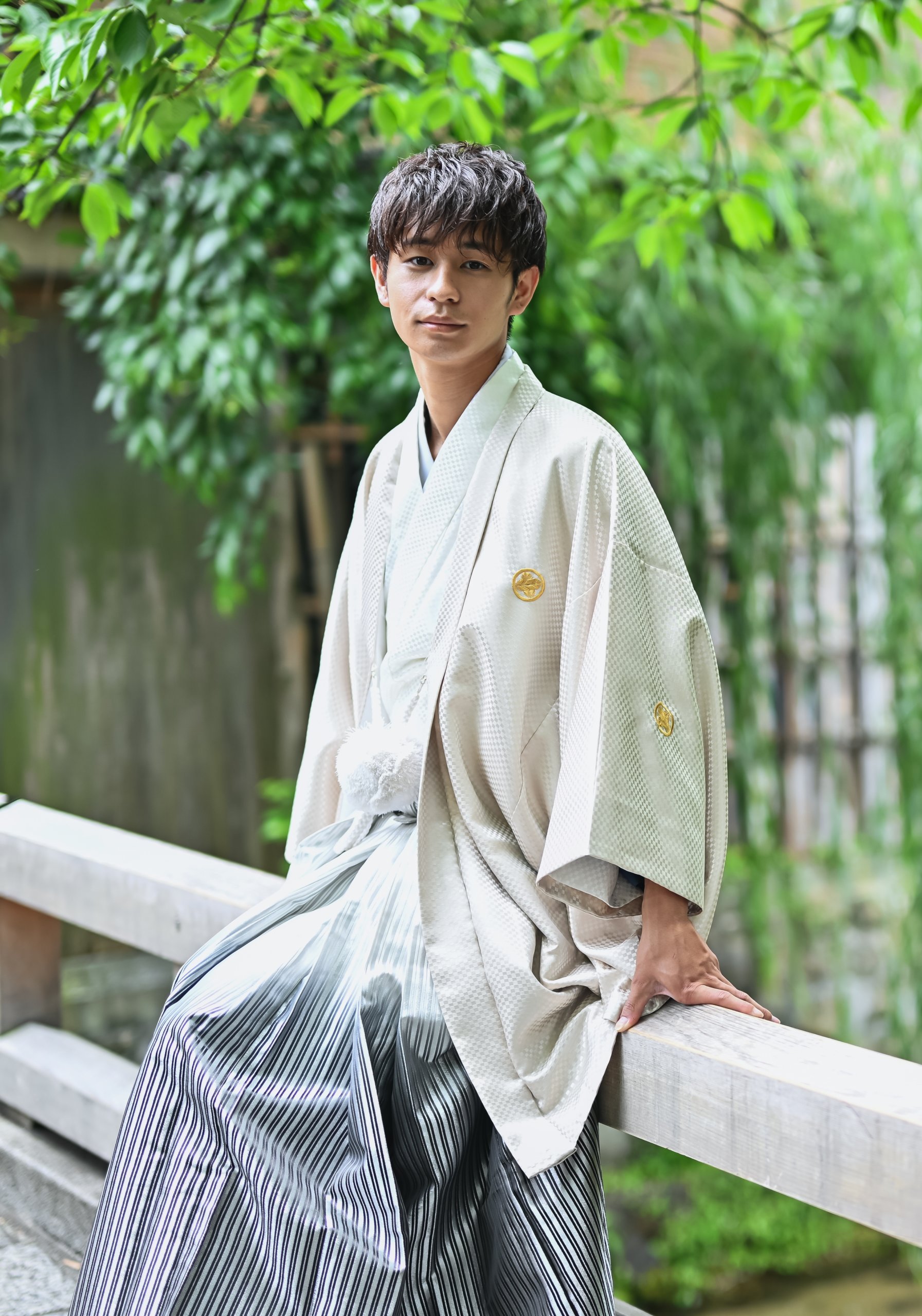 金の家紋があしらわれた羽織袴姿の新郎、緑の植栽を背景にしたポートレート。