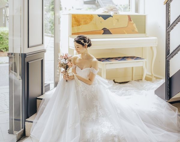 白いアップライトピアノのある明るいエントランスで、白いドレスの新婦がブーケを見つめる。