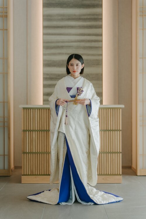 和モダンなスタジオで扇子を持ち正面に立つ白無垢の花嫁の全身ショット。青い掛下と裾のふきが効いた白無垢AR102は前撮りの衣装選びにおすすめ