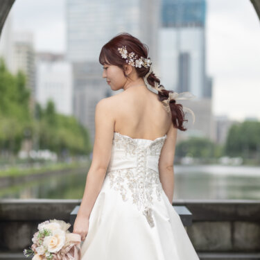 アーチ越しに丸の内の高層ビルを望み、ブーケを手に振り返る花嫁のバックスタイル。ウェディングドレスの編み込みヘアが映える前撮り