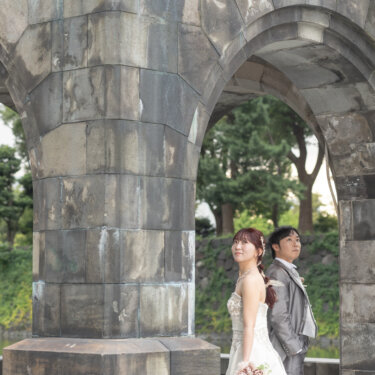 石造りの柱の間からのぞく、ブーケを持ったウェディングドレスの花嫁とグレーのタキシードの新郎。石造りの門で撮るロケーションフォト