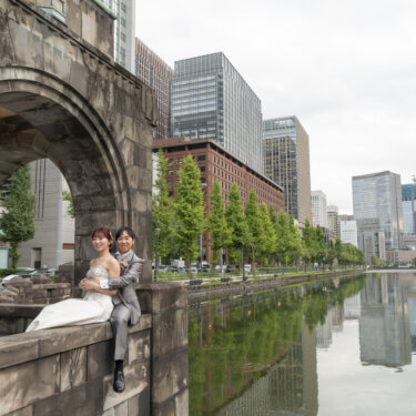 丸の内のビル群が水面に映るお濠のほとりで、石垣に腰かけて寄り添う新郎新婦。東京駅近くで撮るリフレクションの洋装前撮り