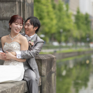 お濠沿いの並木を背に、後ろからハグされて笑い合うタキシードの新郎とウェディングドレスの花嫁。洋装フォトウェディング