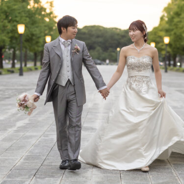 銀杏並木の行幸通りを、手をつないで笑顔で歩くグレーのタキシードの新郎とドレスの裾を持つ花嫁。東京駅前で撮る洋装前撮り