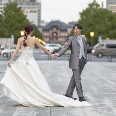 東京駅丸の内駅舎へ向かって、ドレスの裾を持ちながら新郎の手を引く花嫁の後ろ姿。行幸通りで撮るウェディングドレスの前撮り
