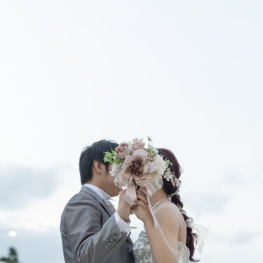 空を背景にピンクのブーケで顔を隠してキスをする新郎とウェディングドレスの花嫁。洋装前撮りの顔隠しポーズ