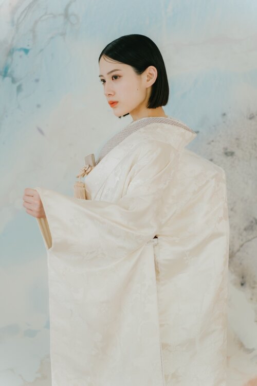 淡いブルーの壁画の前で袖を広げ、横顔を見せる白無垢姿の花嫁。桜の地紋が浮かぶ白無垢AS104は和装前撮りやスタジオフォトで人気の一着