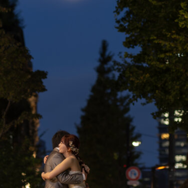 月が浮かぶ夕暮れの並木道で、強く抱き合うタキシードの新郎とウェディングドレスの花嫁。マジックアワーの洋装前撮り