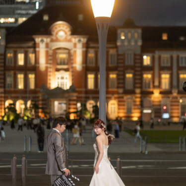 街灯に照らされた夜の東京駅丸の内駅舎の前で、カチンコを持つ新郎とトレーンを引く花嫁。映画風に撮る東京駅の洋装前撮り
