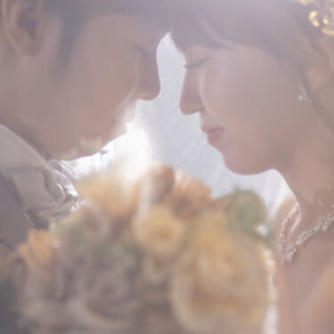 やわらかな逆光の中、ブーケを持っておでこを寄せ合う新郎とウェディングドレスの花嫁の横顔。エモーショナルな前撮り