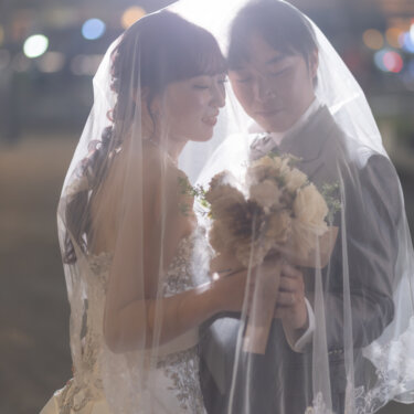 夜の街の光を背に、ベールをかぶって寄り添いブーケを持つ新郎とウェディングドレスの花嫁。東京駅で撮るベールショットの前撮り