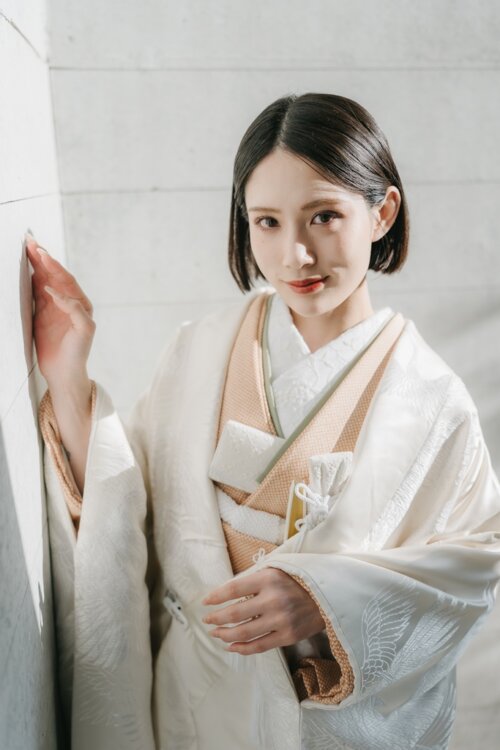 白い壁に手を添えてこちらを見つめる、ベージュの掛下を合わせた白無垢の花嫁。鶴の地紋が映えるスタジオフォト