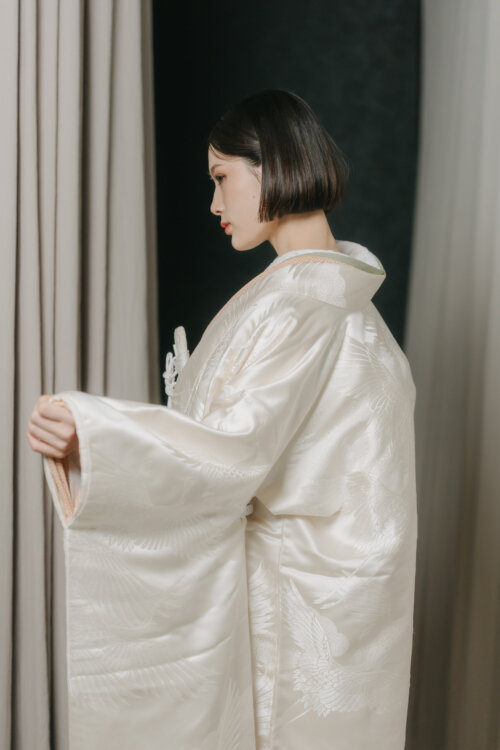 カーテンの前で袖を広げて横顔を見せる白無垢の花嫁。鶴の織り柄が浮かぶ衣装の質感が分かる、スタジオフォトの衣装カット