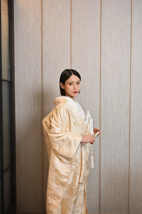 木目の壁の前で扇子と房飾りを持ち、正面を向いて立つ金糸の刺繍入りの白無垢の花嫁。スタジオフォトで撮影した衣装写真