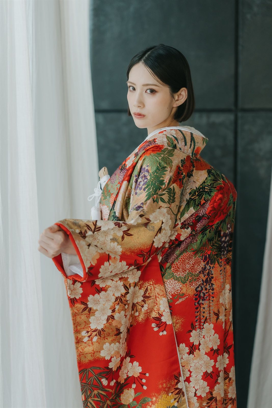 白いカーテン越しの柔らかな光の中で振り返る、赤地に桜と牡丹を織り出した色打掛の花嫁。和装前撮りで人気のモダンな色打掛コーディネート